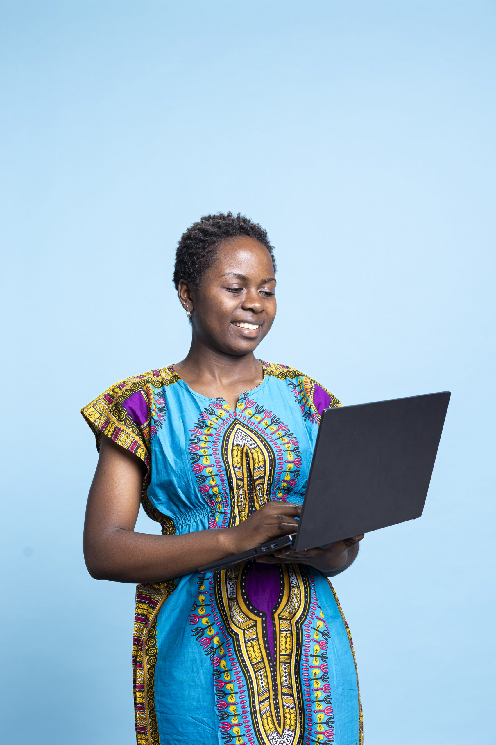 Young online user works on notebook computer to access internet sites while exploring social media in a studio. African american girl smiles for the camera while she is holding electronic device.